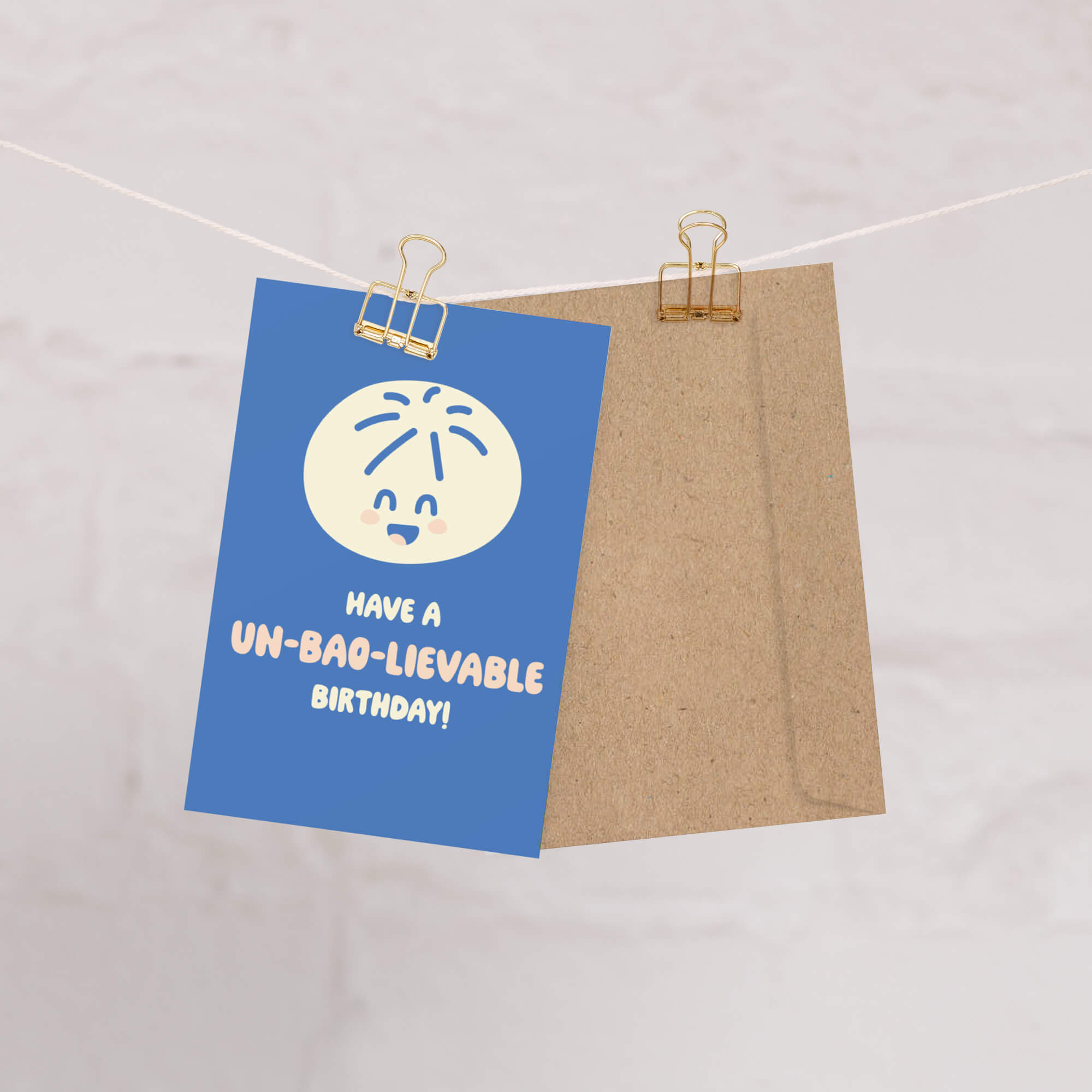Blue card with a cartoon face and 'Have a un-bag-lievable birthday!' text, attached to a brown bag with a clip on a white background.