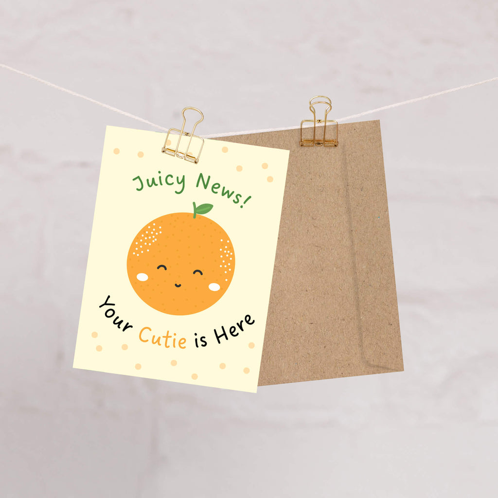 A cute new baby card and kraft envelope hanging from a string with two binder clips. The card is cream-colored with a smiling orange illustration and the text 'Juicy News! Your Cutie is Here'.