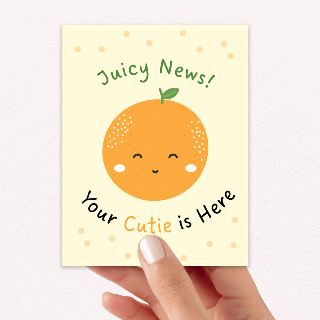 A hand holding a new baby shower greeting card with a cute orange illustration and the text 'Juicy News! Your Cutie is Here'.