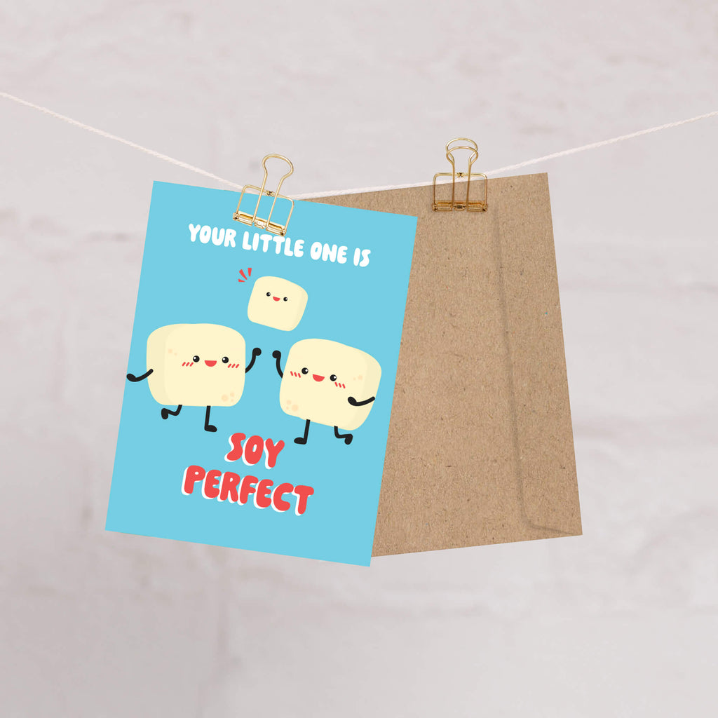 A cute baby shower card with a light blue background and cartoon tofus, clipped to a string with a kraft paper envelope behind it on a light marble-like surface.