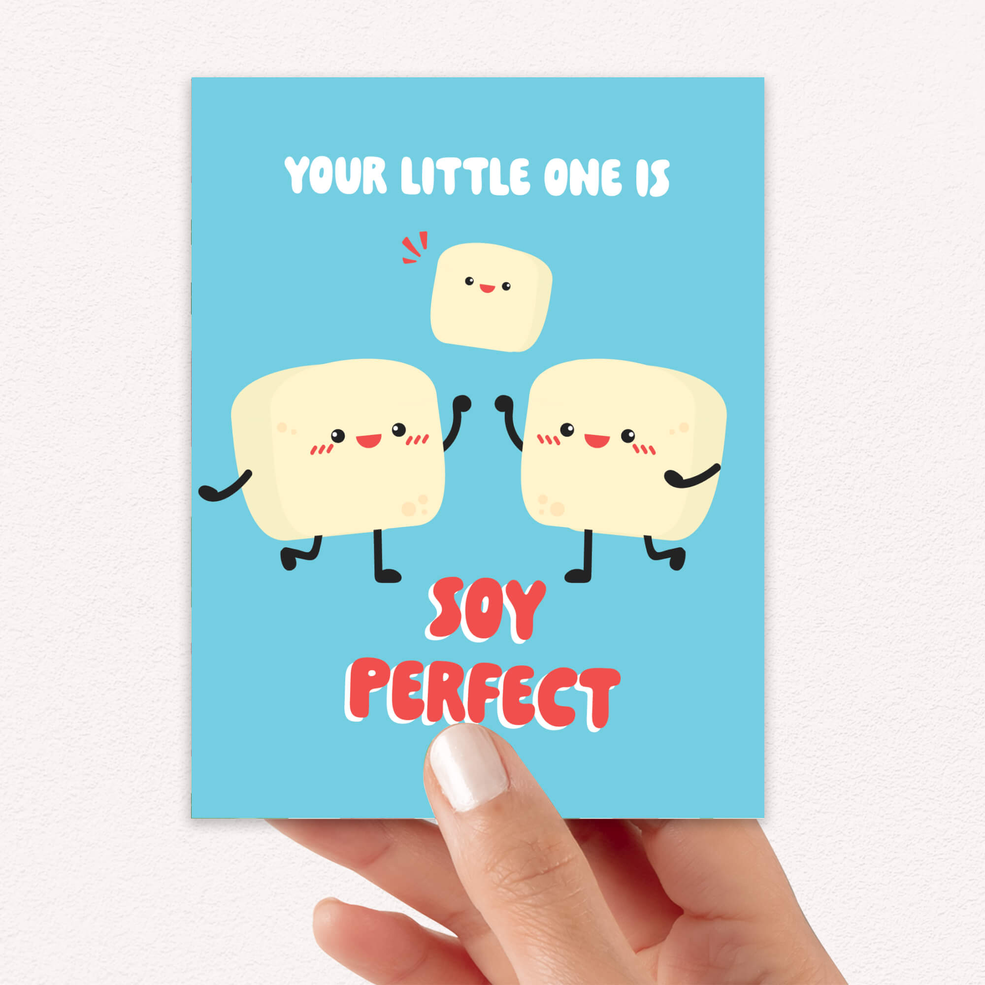 A hand holds a baby shower card with a cute cartoon illustration of three marshmallows and the text 'YOUR LITTLE ONE IS SOY PERFECT'.