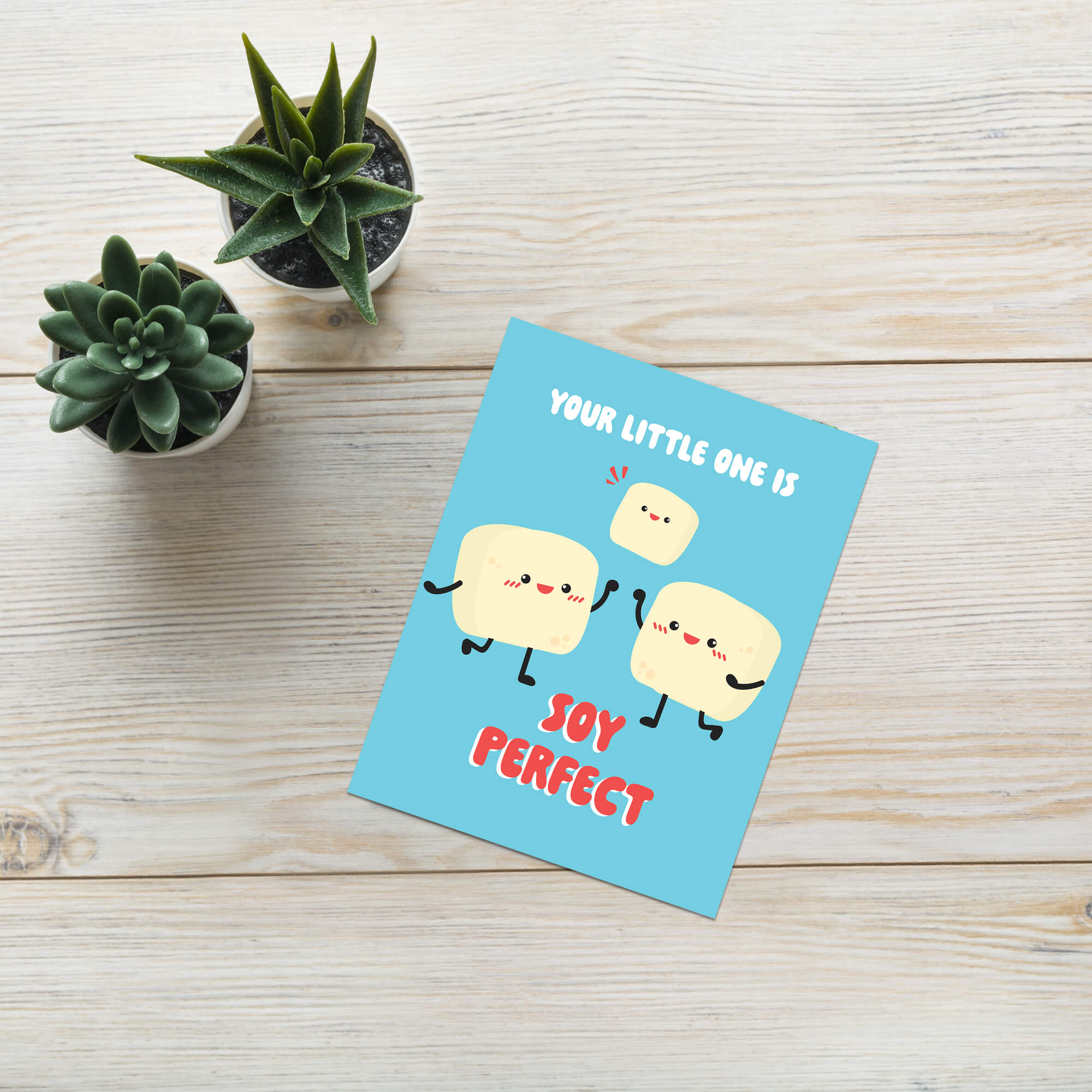 A funny baby shower card with a cute cartoon design of three smiling cream-colored tofus and the text 'YOUR LITTLE ONE IS SOY PERFECT' lies on a light wood surface next to two small potted succulents.