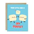 A funny baby shower card with a light blue front and a kraft envelope behind it. The card features a cute cartoon illustration of three smiling tofus holding hands, with the text 'YOUR LITTLE ONE IS' above and 'SOY PERFECT' below in red and white lettering.