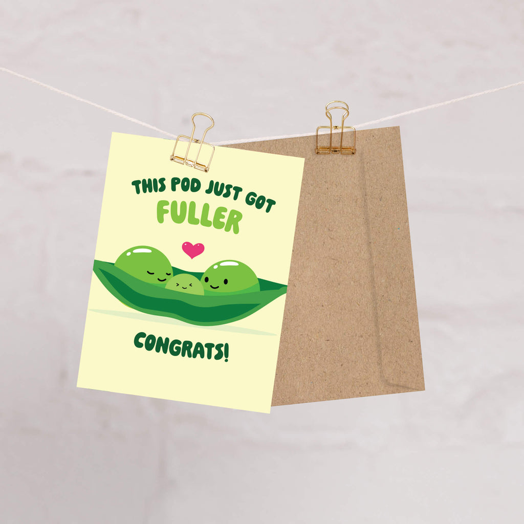 A novelty greeting card with a cartoon pea pod illustration and the text 'THIS POD JUST GOT FULLER CONGRATS!' is clipped to a string with a kraft paper envelope behind it on a light marble-like background.