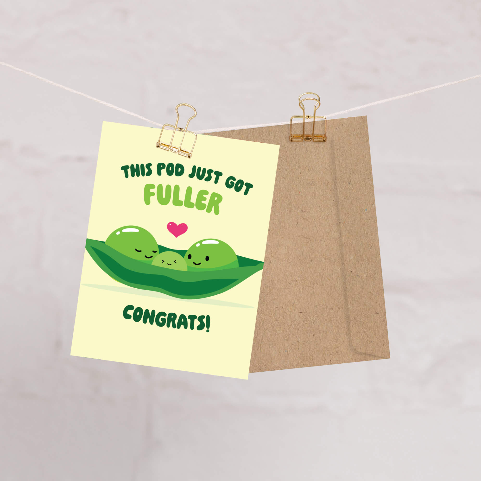 A novelty greeting card with a cartoon pea pod illustration and the text 'THIS POD JUST GOT FULLER CONGRATS!' is clipped to a string with a kraft paper envelope behind it on a light marble-like background.