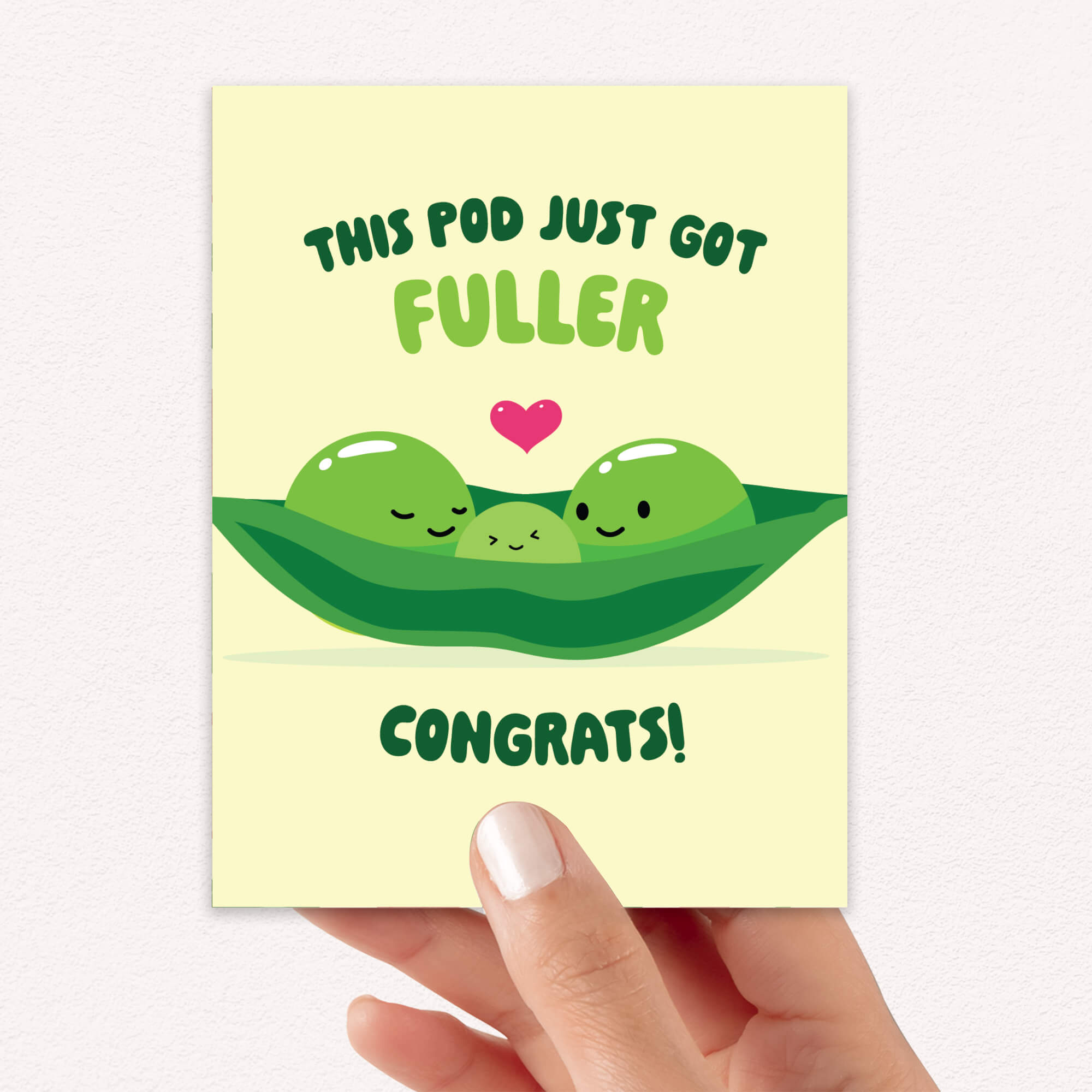 A hand holding a pale yellow baby shower card with a cute cartoon illustration of three green peas in a pod and the text 'THIS POD JUST GOT FULLER CONGRATS!'.