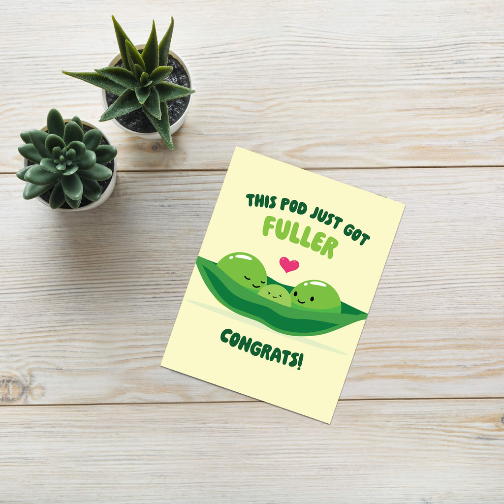 A flat-lay photograph of a yellow edamame baby shower card with a cute green pea pod illustration and the text 'THIS POD JUST GOT FULLER CONGRATS!'. The card is placed on a light-colored wooden surface next to two small potted succulents.