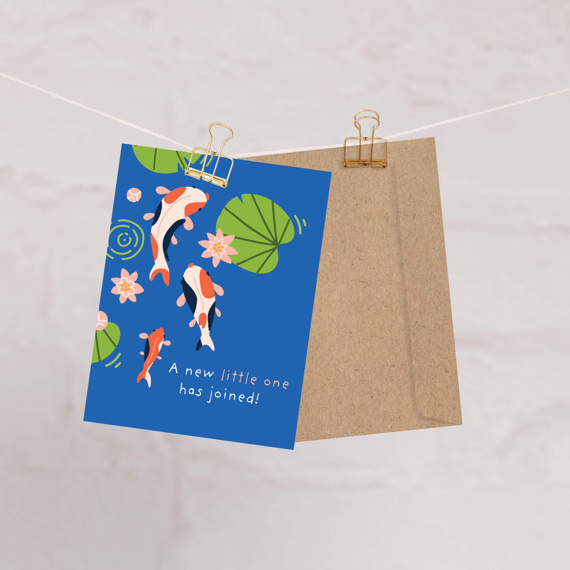 A baby shower card with illustrated koi fish, lily pads, and pink flowers hangs from a string with two gold binder clips, shown with a brown kraft envelope behind it on a light marble-like background.