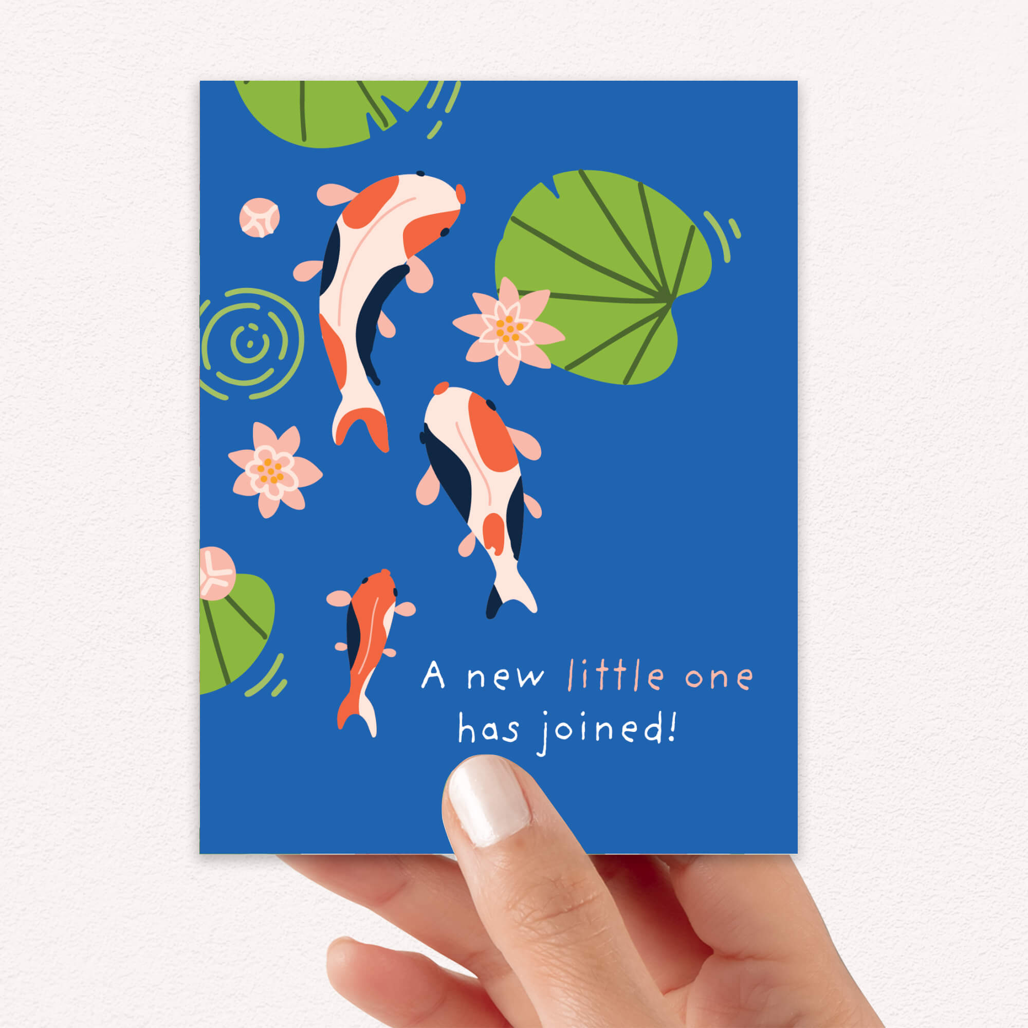 A hand holding a blue new baby card with illustrations of koi fish, lily pads, and pink water lilies, with the text 'A new little one has joined!'.