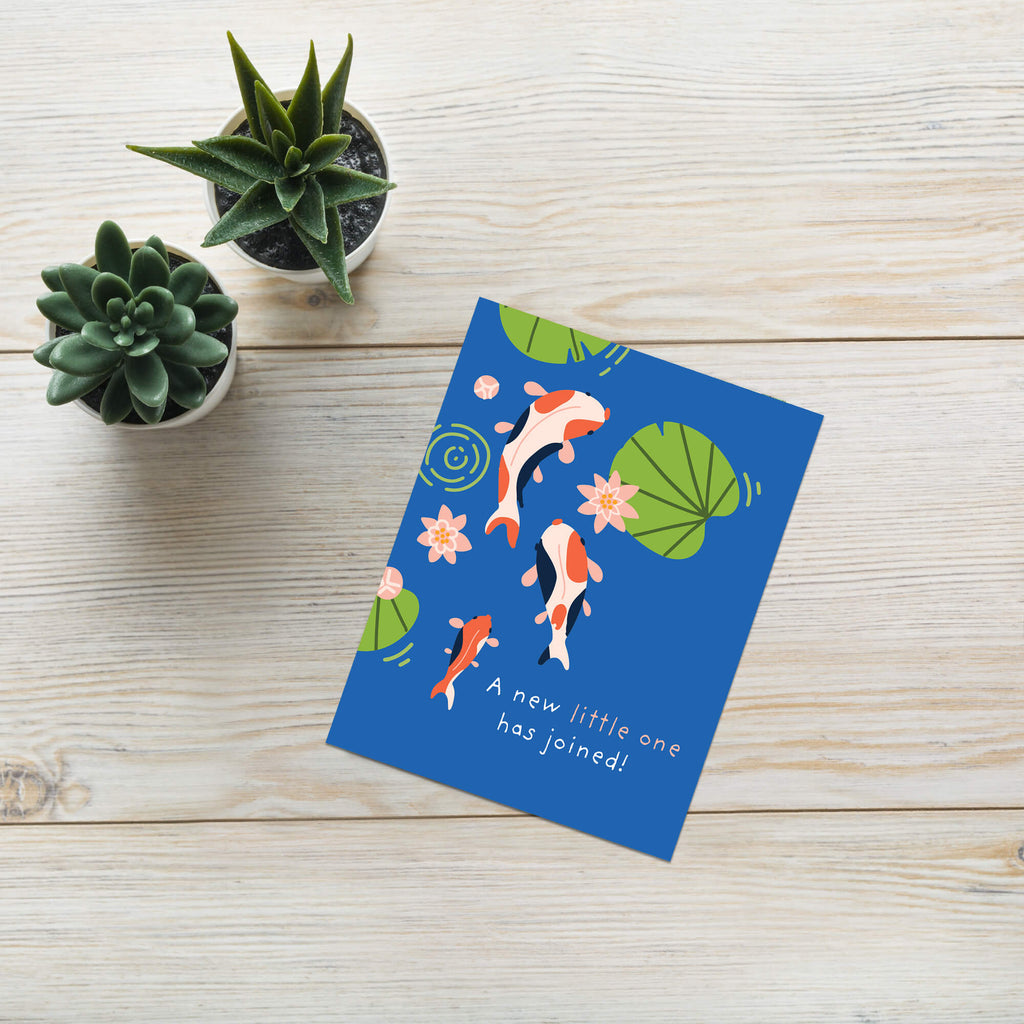 A blue baby shower card featuring koi fish, lily pads, and the text 'A new little one has joined!' in white, styled on a light wood background next to three small potted succulents.