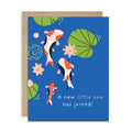 A Japanese baby shower card with a blue background featuring illustrated koi fish, lily pads, and pink water lilies, with the text 'A new little one has joined!' in white. A kraft paper envelope is placed behind the card on a white background.