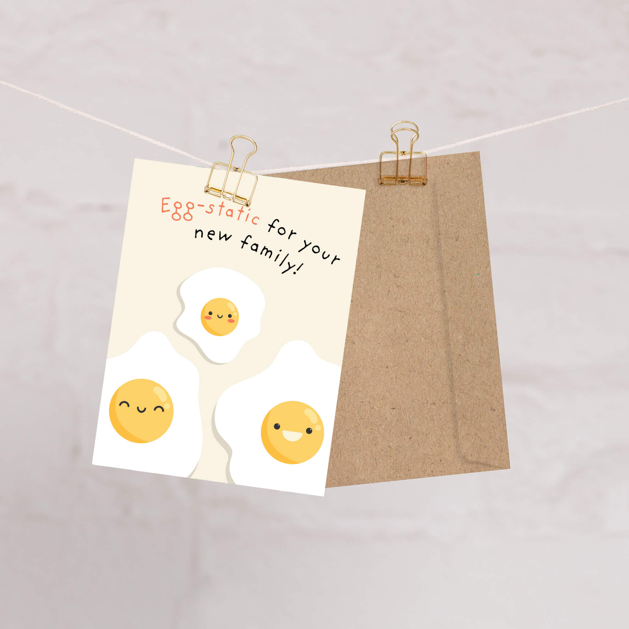 A funny baby shower card with a kraft envelope, clipped to a string against a light marble background. The card reads 'Egg-static for your new family!' and features cute cartoon fried eggs.