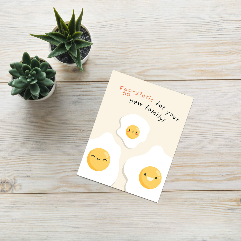 A funny baby shower card styled on a light wood tabletop next to two small potted succulents. The card features a cute illustration of three smiling fried eggs on a cream background with the text 'Egg-static for your new family!'.