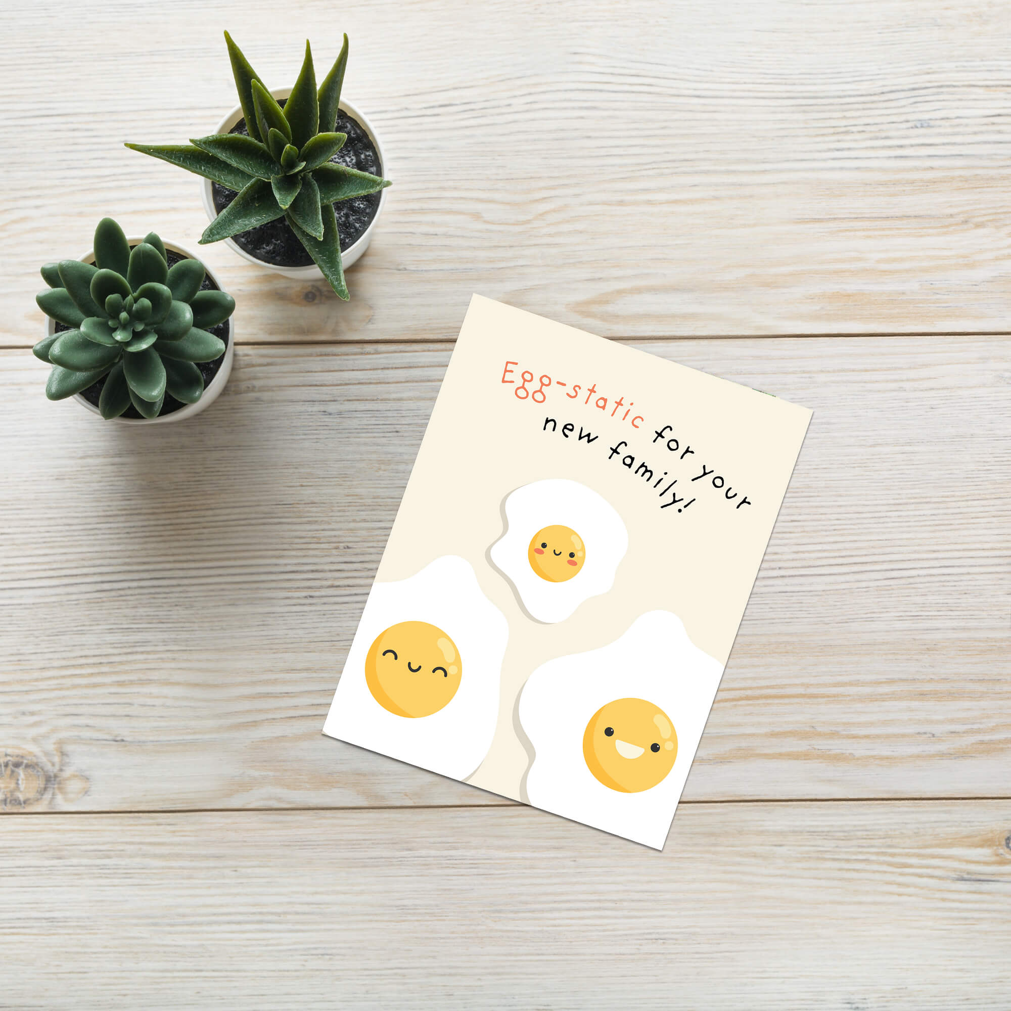 A funny baby shower card styled on a light wood tabletop next to two small potted succulents. The card features a cute illustration of three smiling fried eggs on a cream background with the text 'Egg-static for your new family!'.