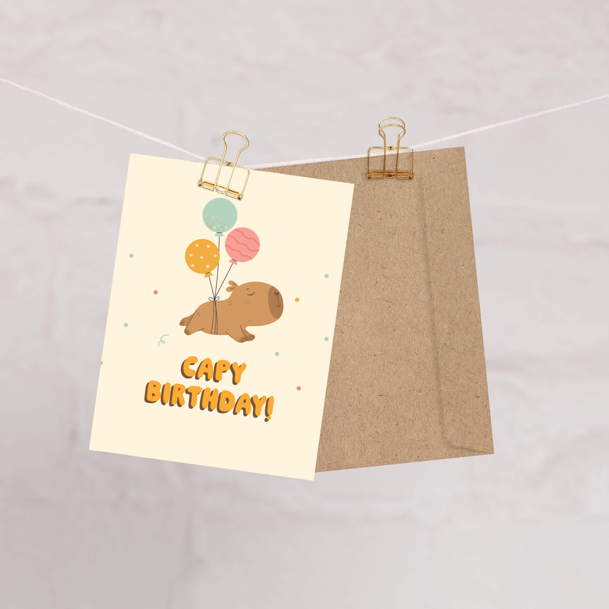 A cute capybara birthday card with balloons and the text 'Capy Birthday!' hanging from a string with two gold binder clips, shown with a brown kraft envelope behind it on a light marble background.