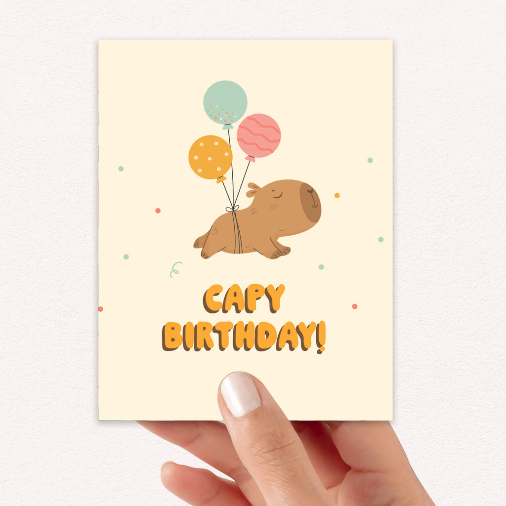 A hand holding a capybara birthday card with a cream background, featuring a cartoon capybara floating with three balloons and the text 'CAPY BIRTHDAY!' in orange. The card is shown against a light textured wall.