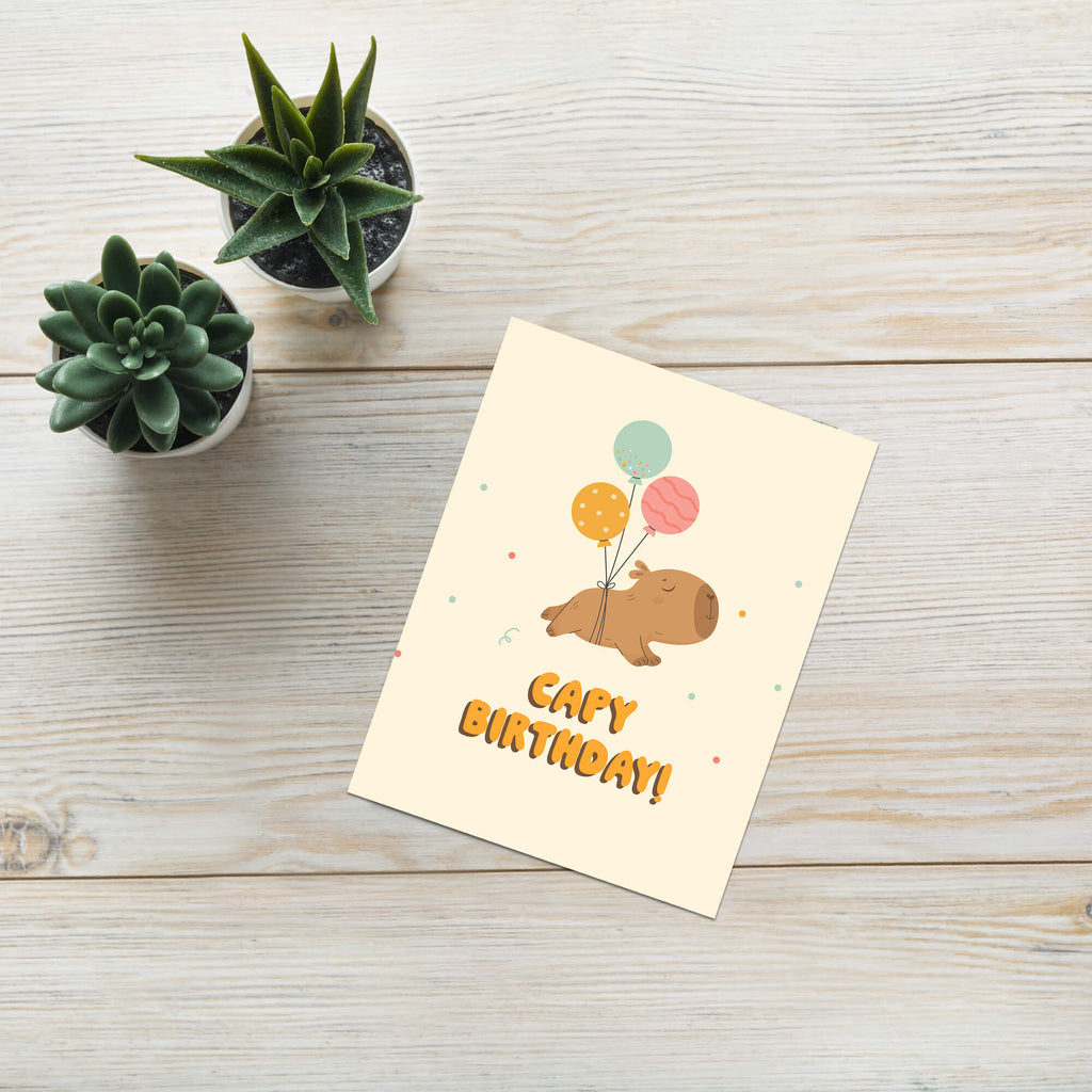 A capybara birthday card with a cute cartoon pig holding balloons, placed on a light wood surface next to two small potted succulents. The card reads 'Cute Birthday!'.