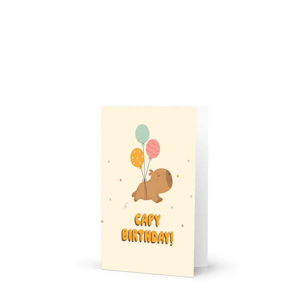 A capybara birthday card with a cream background featuring a cartoon capybara holding balloons and the text 'Capy Birthday!' in orange, shown standing upright on a white background.