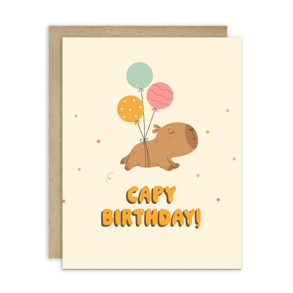 A cute capybara birthday card with a kraft envelope. The card has a cream background, featuring a cartoon capybara holding three balloons (mint green, orange, and pink) with the text 'CAPY BIRTHDAY!' in orange.