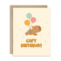 A cute capybara birthday card with a kraft envelope. The card has a cream background, featuring a cartoon capybara holding three balloons (mint green, orange, and pink) with the text 'CAPY BIRTHDAY!' in orange.