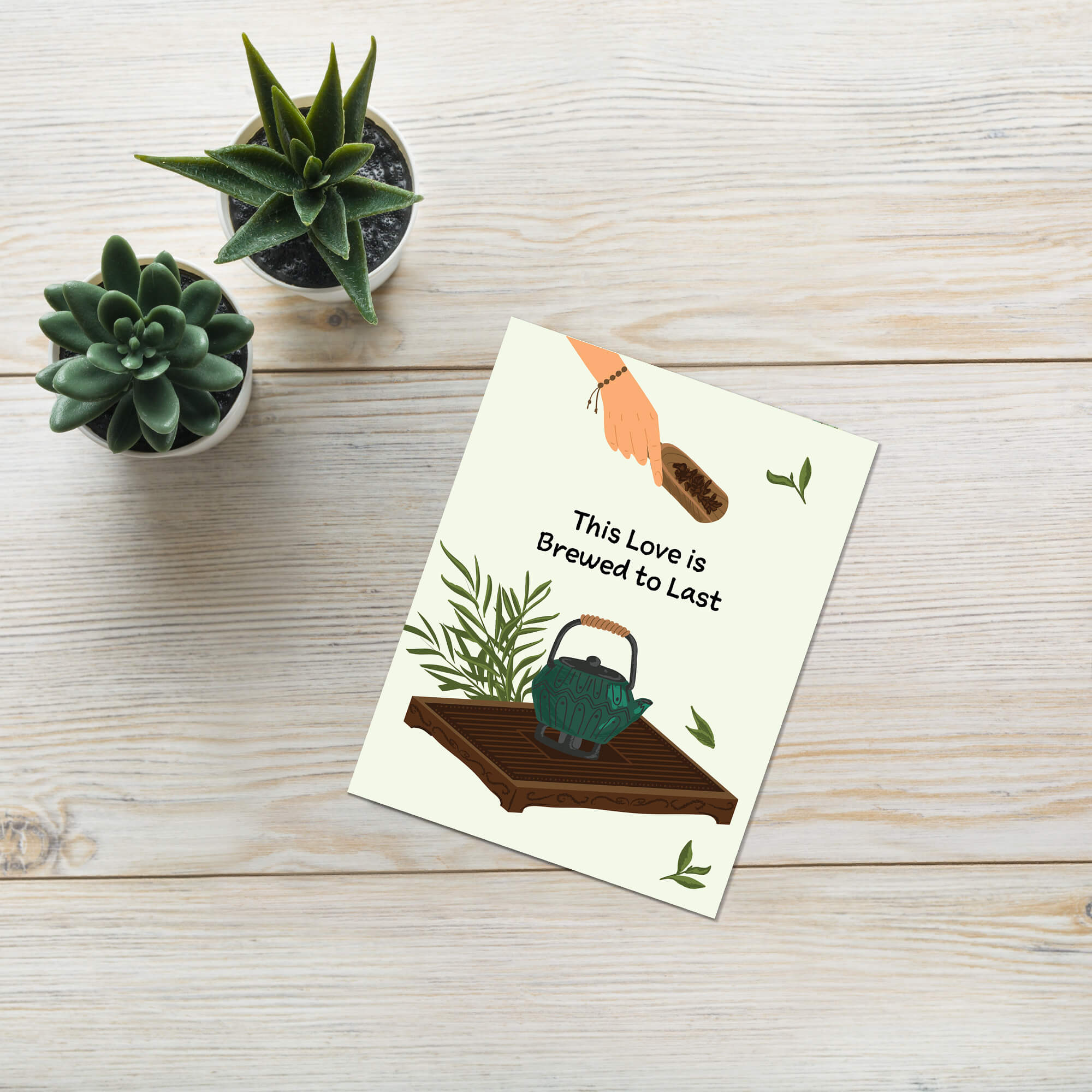 Greeting card with teapot illustration and text 'This Love is Brewed to Last' on a wooden surface with potted plants.