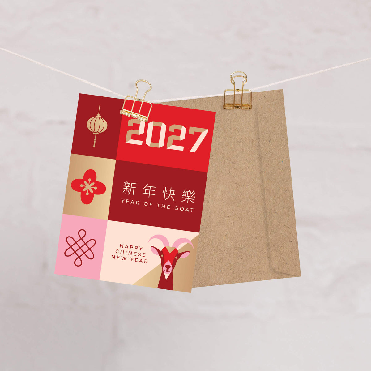 A square Chinese New Year greeting card clipped to a string with a kraft paper envelope behind it. The card features a red, beige, and pink grid design with goat illustrations, the year 2027, and Chinese and English New Year text.