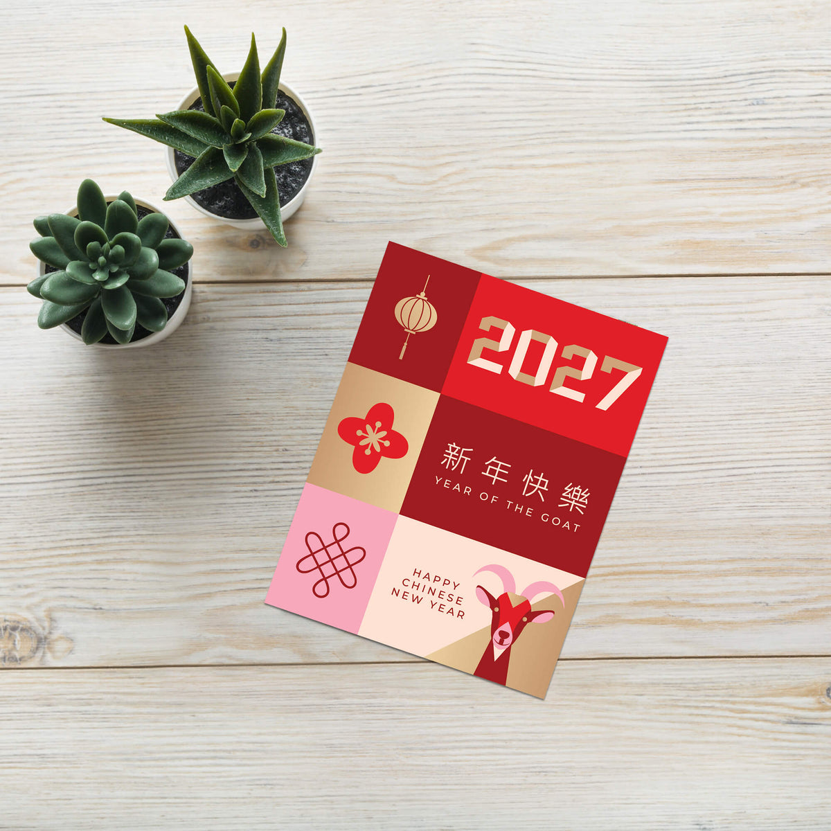 A 2027 Chinese New Year greeting card featuring red, gold, and pink panels with goat motifs and Chinese text, styled on a light wood surface next to two small potted succulents.