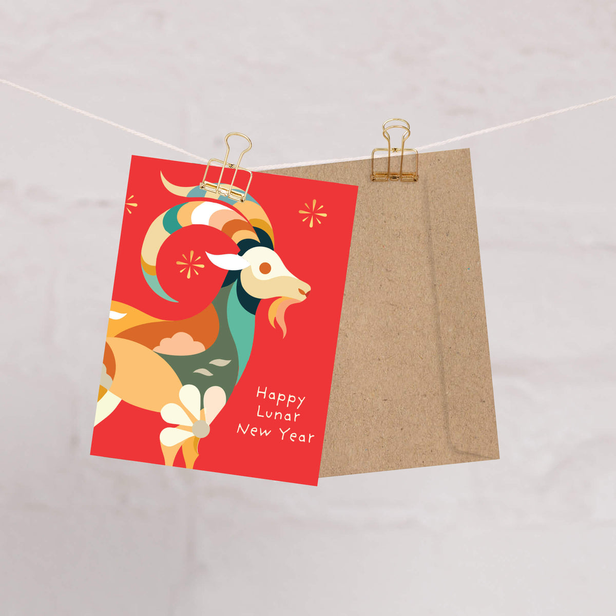 A red chinese new year card with a stylized ram illustration and the words 'Happy Lunar New Year' is clipped to a string next to a kraft paper envelope on a light marble-like background.