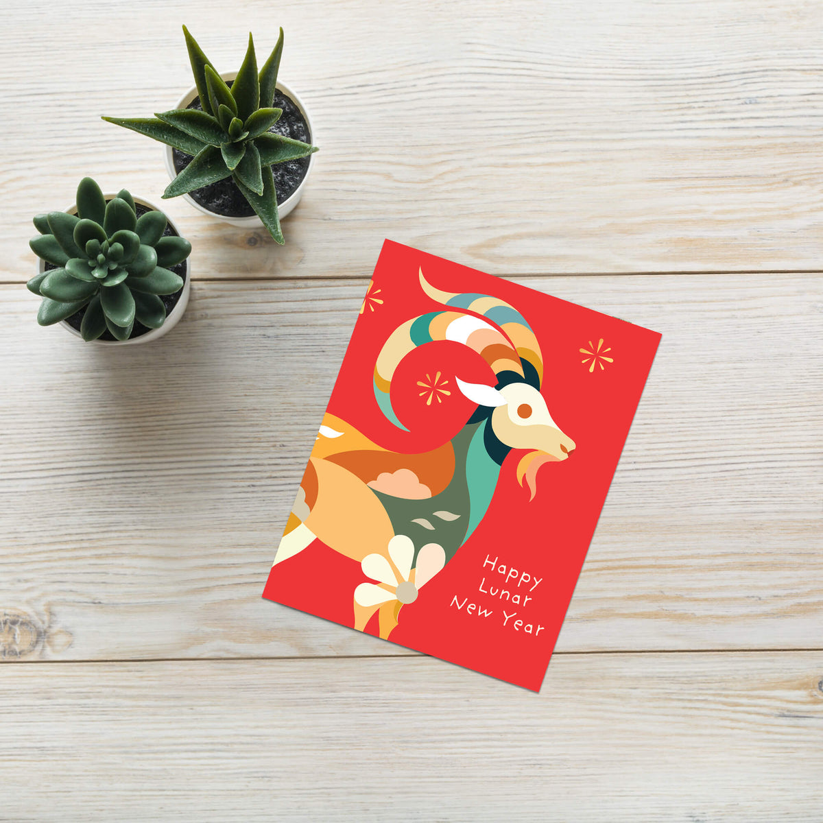 A red chinese new year card 2027 featuring a stylized ram illustration and the text 'Happy Lunar New Year' in white, placed on a light wood surface next to two small potted succulents.