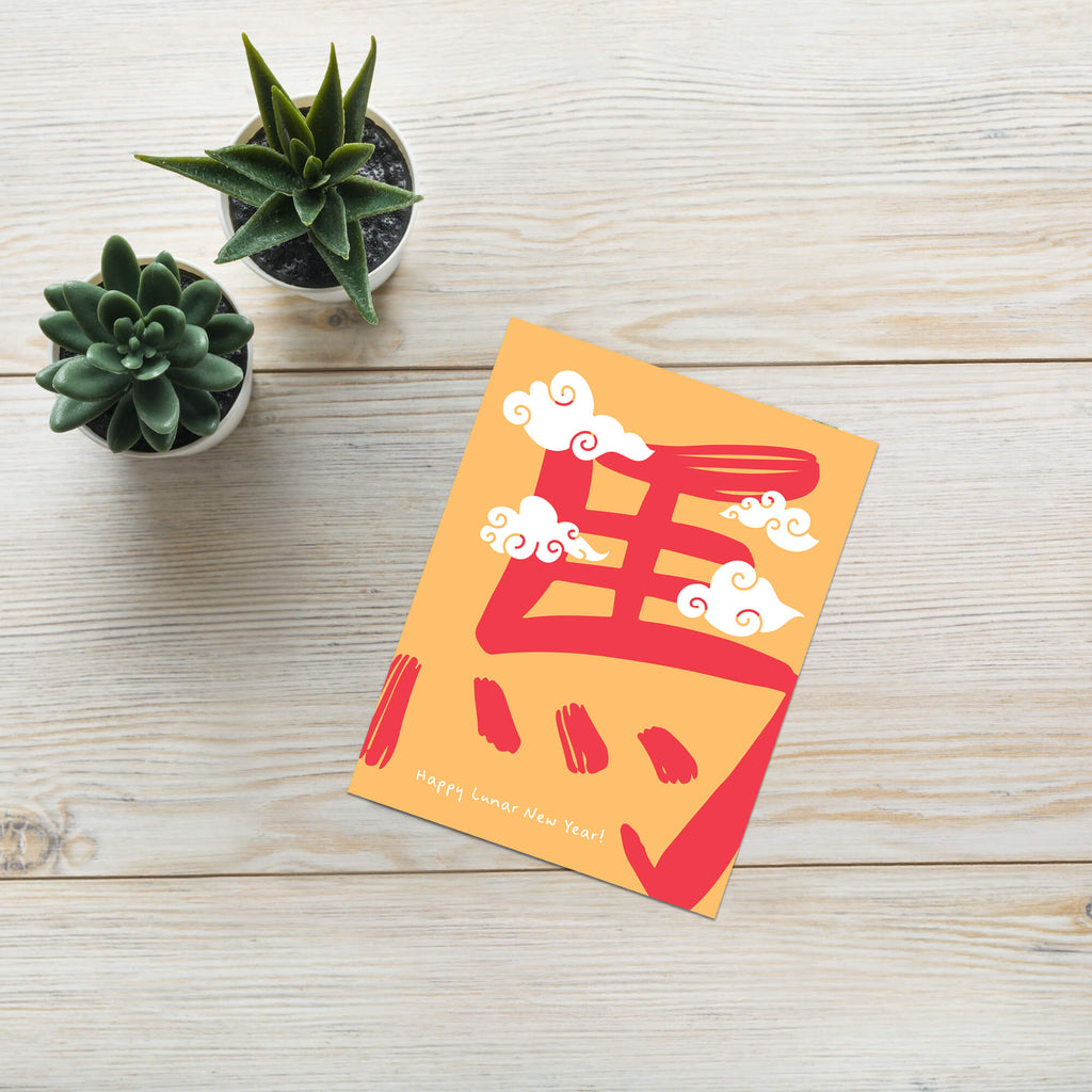 Orange card with red and white design on a wooden surface with two small potted plants.