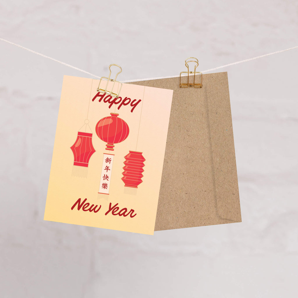 A lunar New Year greeting card with red lantern illustrations and the words 'Happy New Year' in red script, clipped to a string with a kraft paper envelope behind it against a light textured background.