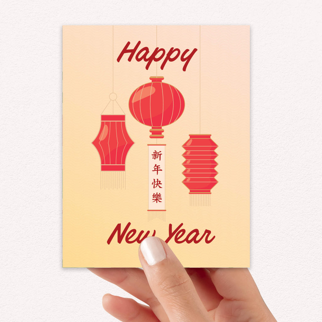A hand holding a chinese new year greeting card with a warm cream background, red hanging lantern illustrations, and the text 'Happy New Year' in red script. The central vertical text includes Chinese characters for a happy new year.