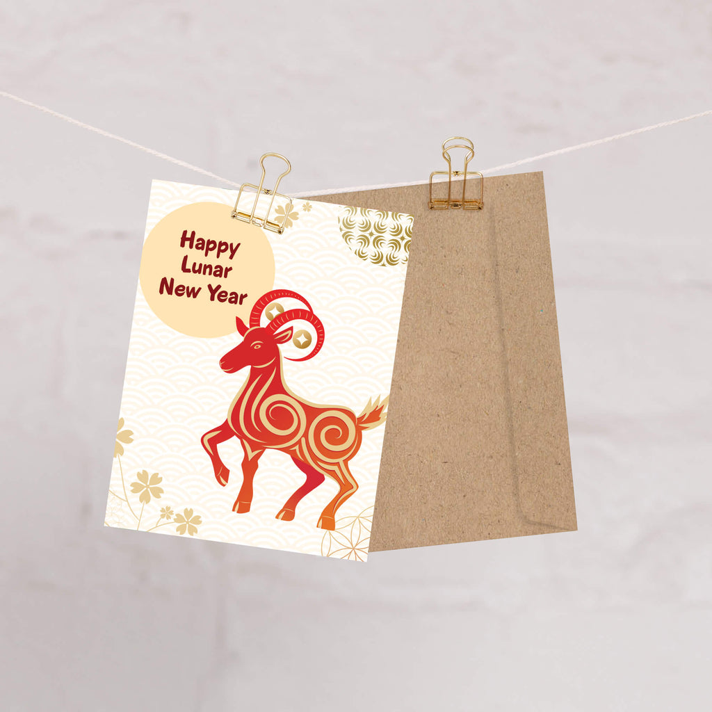 A chinese new year card for the Lunar New Year hangs from a string with two gold binder clips. The card features a red and orange ram illustration and a beige circle reading 'Happy Lunar New Year.