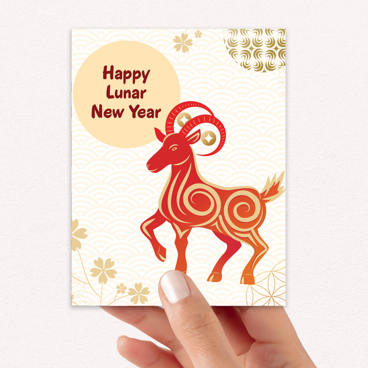 A hand holding a Chinese New Year greeting card 2027 featuring a stylized red and orange ram with gold patterns, a beige circle reading 'Happy Lunar New Year,' and gold floral motifs on a cream background.