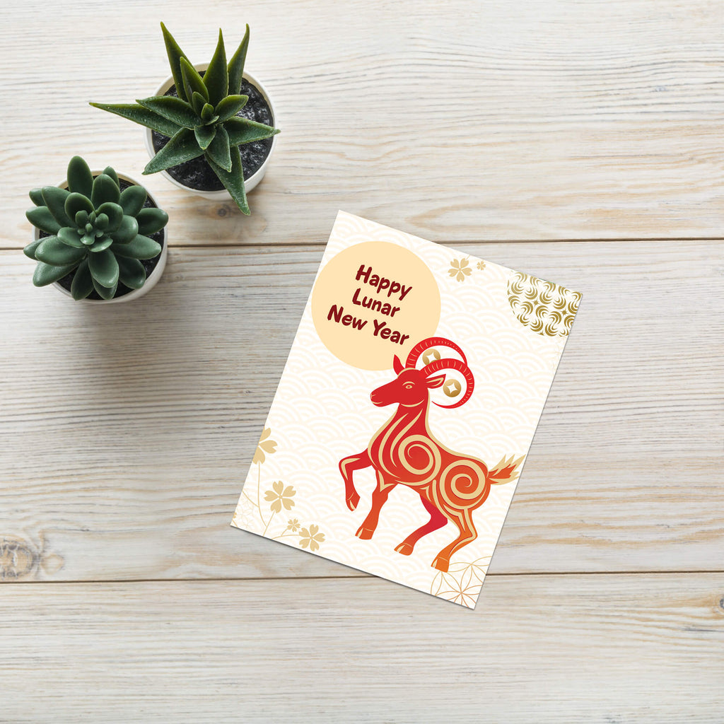 A Year of the Goat card with a stylized red ram illustration and the text 'Happy Lunar New Year' lies on a light wood surface. Two small potted succulents are placed above the card.