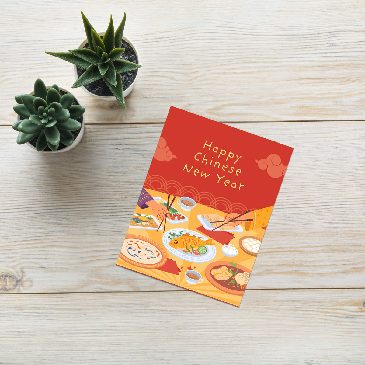 A red and orange Chinese New Year greeting card with 'Happy Chinese New Year' written on it, styled on a light wood tabletop next to two small potted succulents.