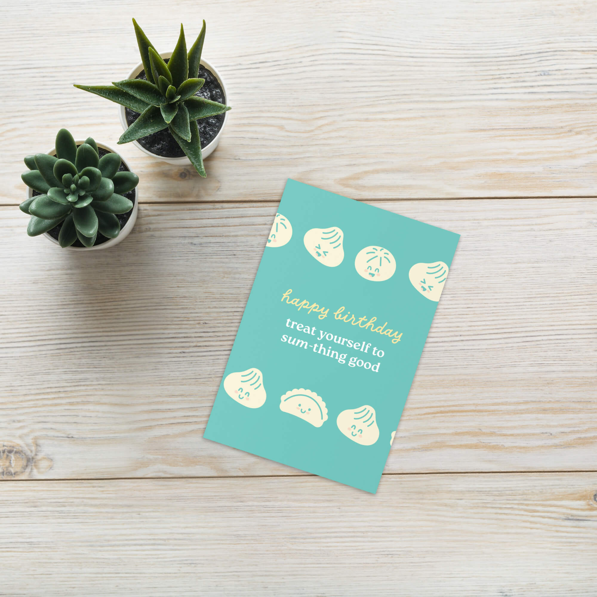 Turquoise birthday card with decorative elements on a wooden surface with small potted plants.