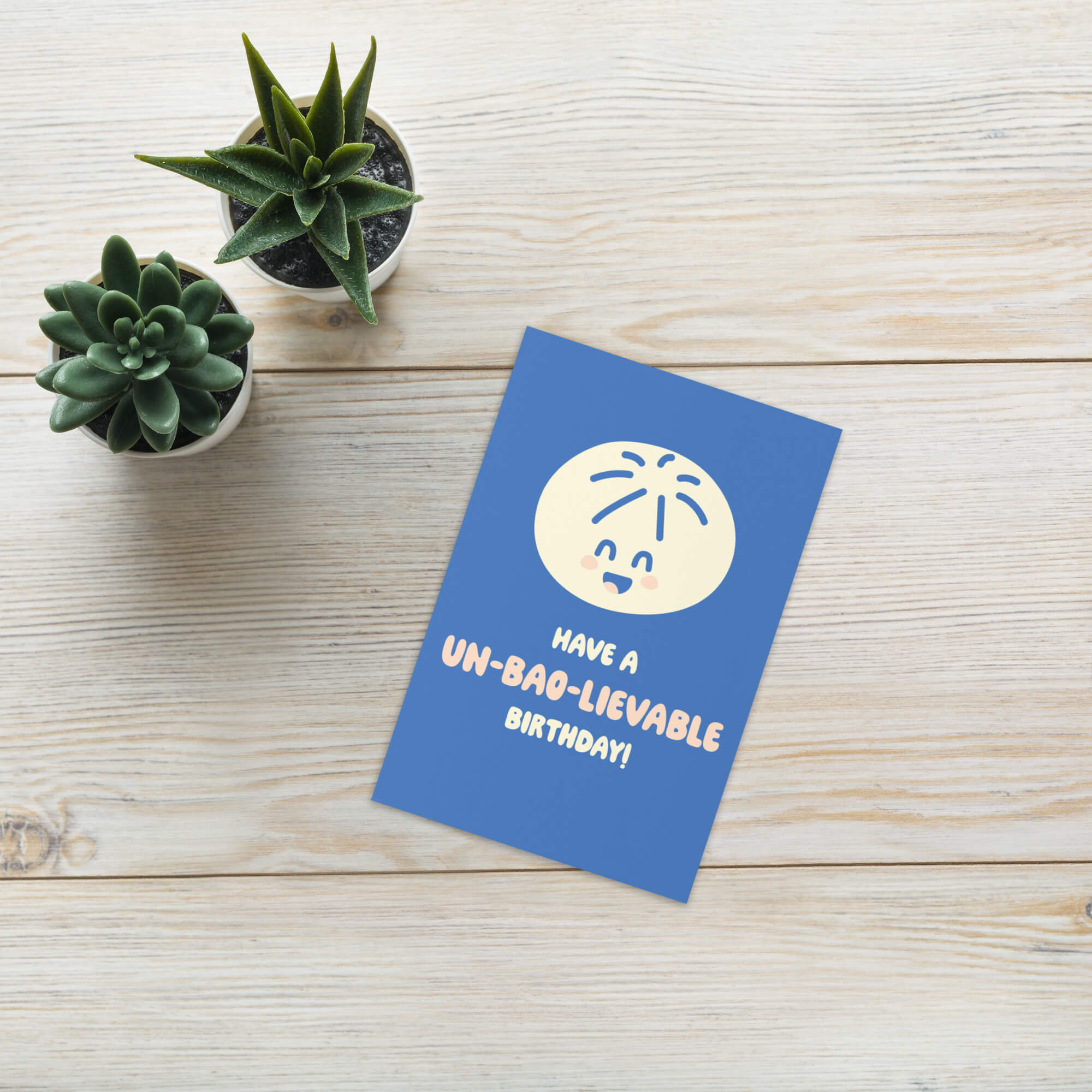 Blue birthday card with a cartoon face and text on a wooden surface with small potted plants.