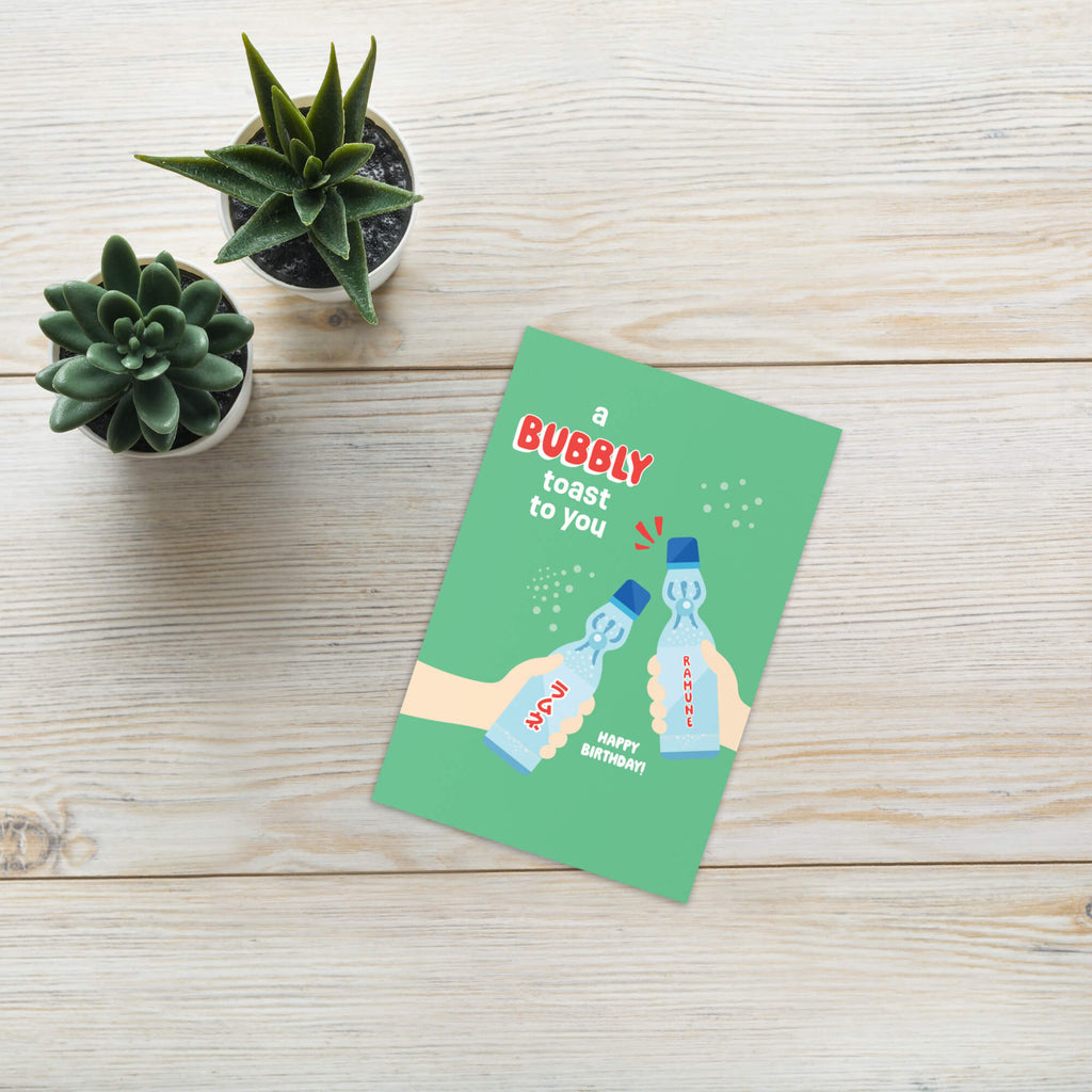 Green birthday card with ramune bottles and text on a wooden surface with small plants.