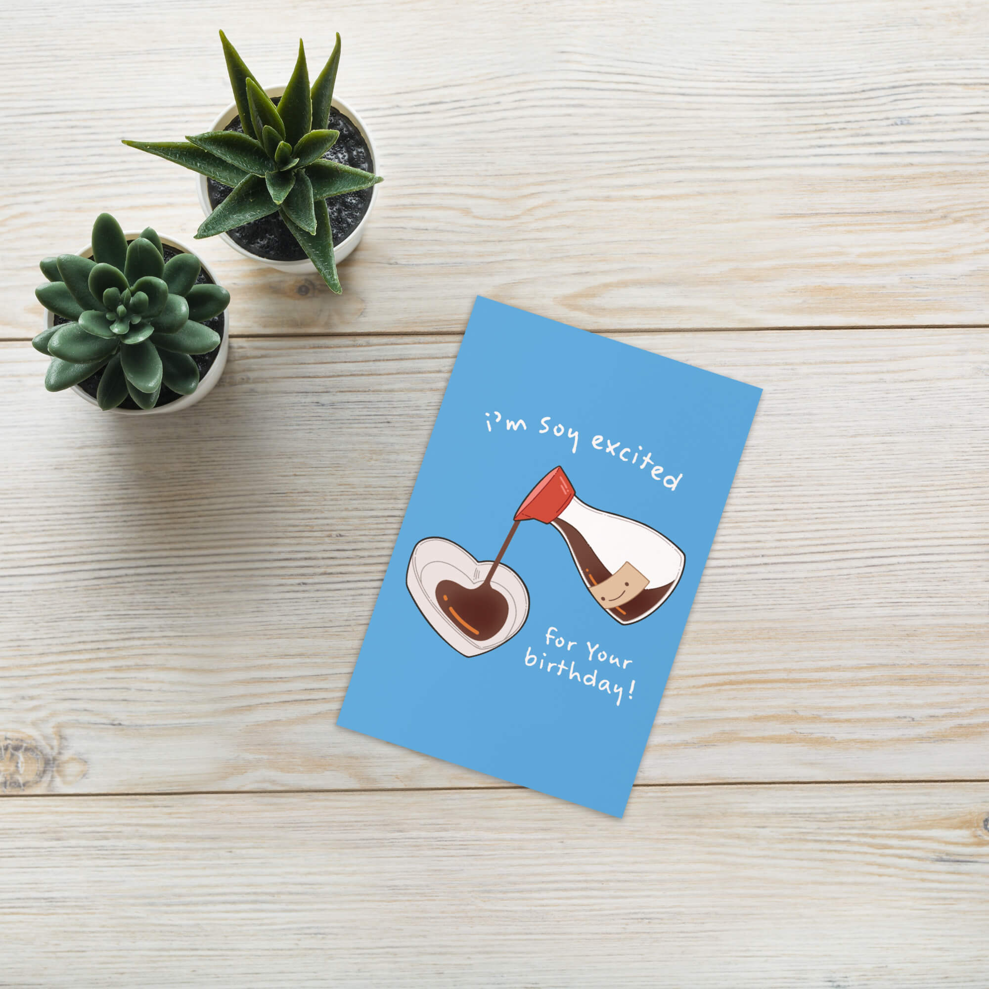 Blue birthday card with a design of a cake being poured into a glass, text 'I'm so excited for your birthday!', on a wooden surface with two small potted plants.