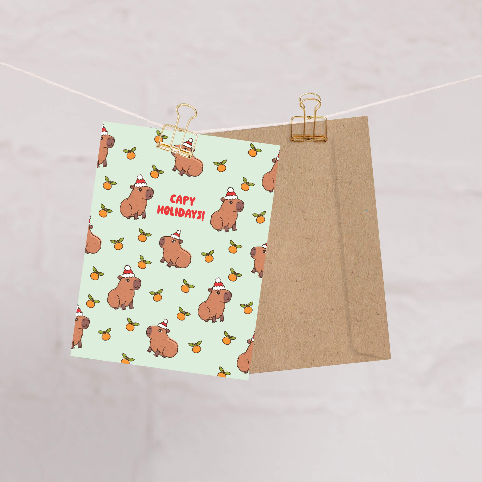 cute christmas card with Repeating pattern of capybaras in Santa hats with small oranges on a pale green background. Text reads a pun: “Capy Holidays.”