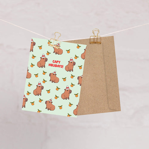 cute christmas card with Repeating pattern of capybaras in Santa hats with small oranges on a pale green background. Text reads a pun: “Capy Holidays.”