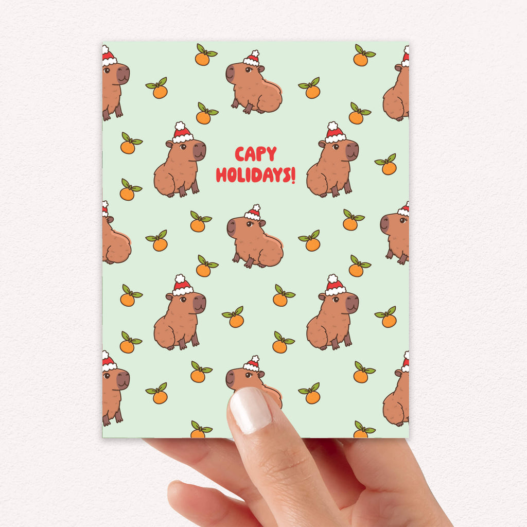 cute christmas card with Repeating pattern of capybaras in Santa hats with small oranges on a pale green background. Text reads a pun: “Capy Holidays.”