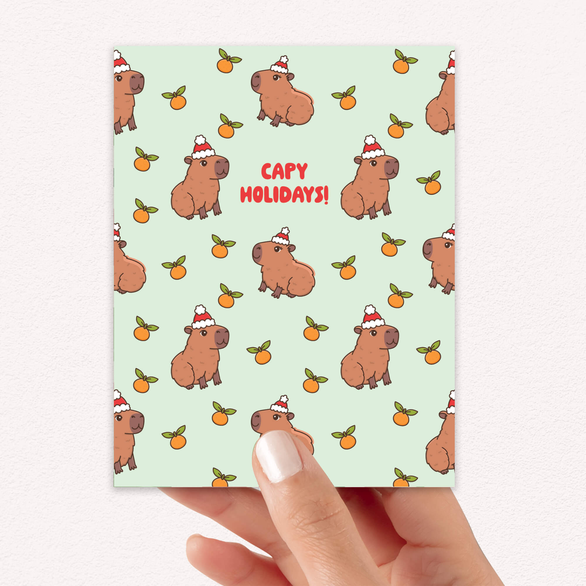 cute christmas card with Repeating pattern of capybaras in Santa hats with small oranges on a pale green background. Text reads a pun: “Capy Holidays.”