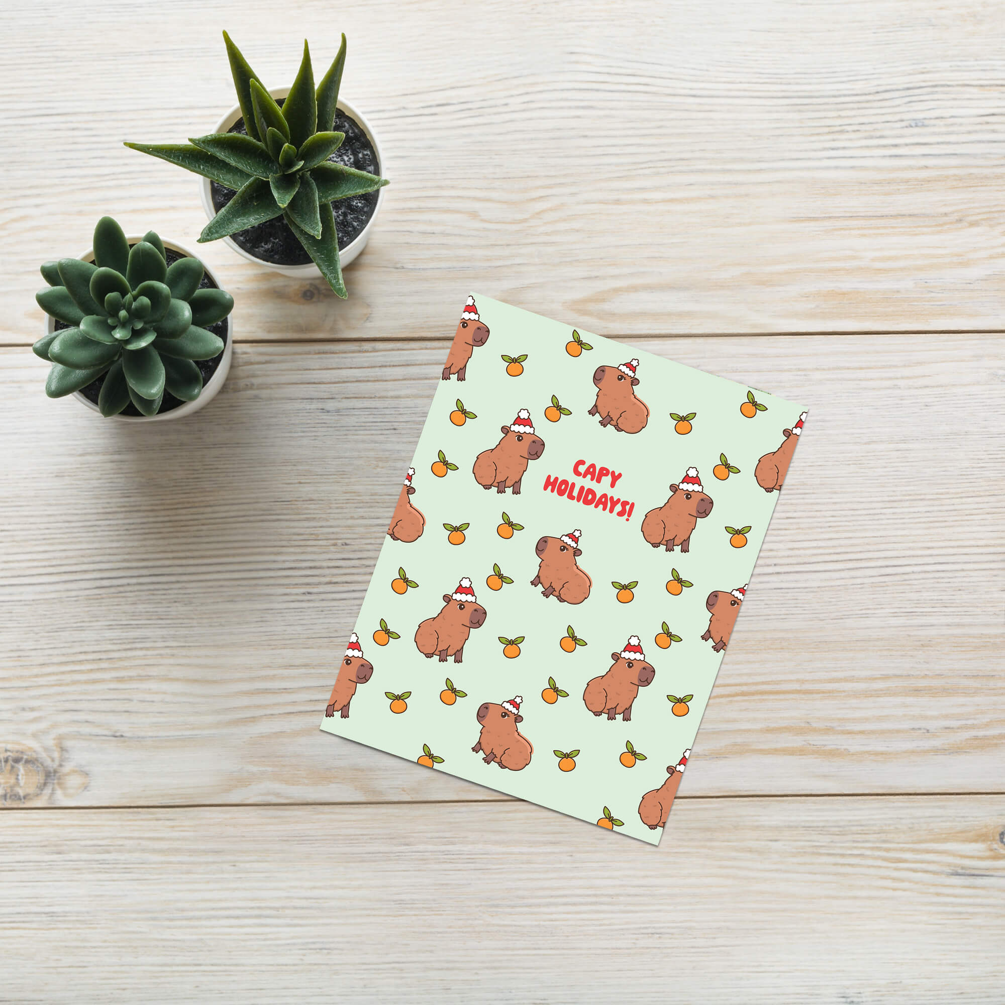 cute christmas card with Repeating pattern of capybaras in Santa hats with small oranges on a pale green background. Text reads a pun: “Capy Holidays.”