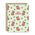 cute christmas card with Repeating pattern of capybaras in Santa hats with small oranges on a pale green background. Text reads a pun: “Capy Holidays.”
