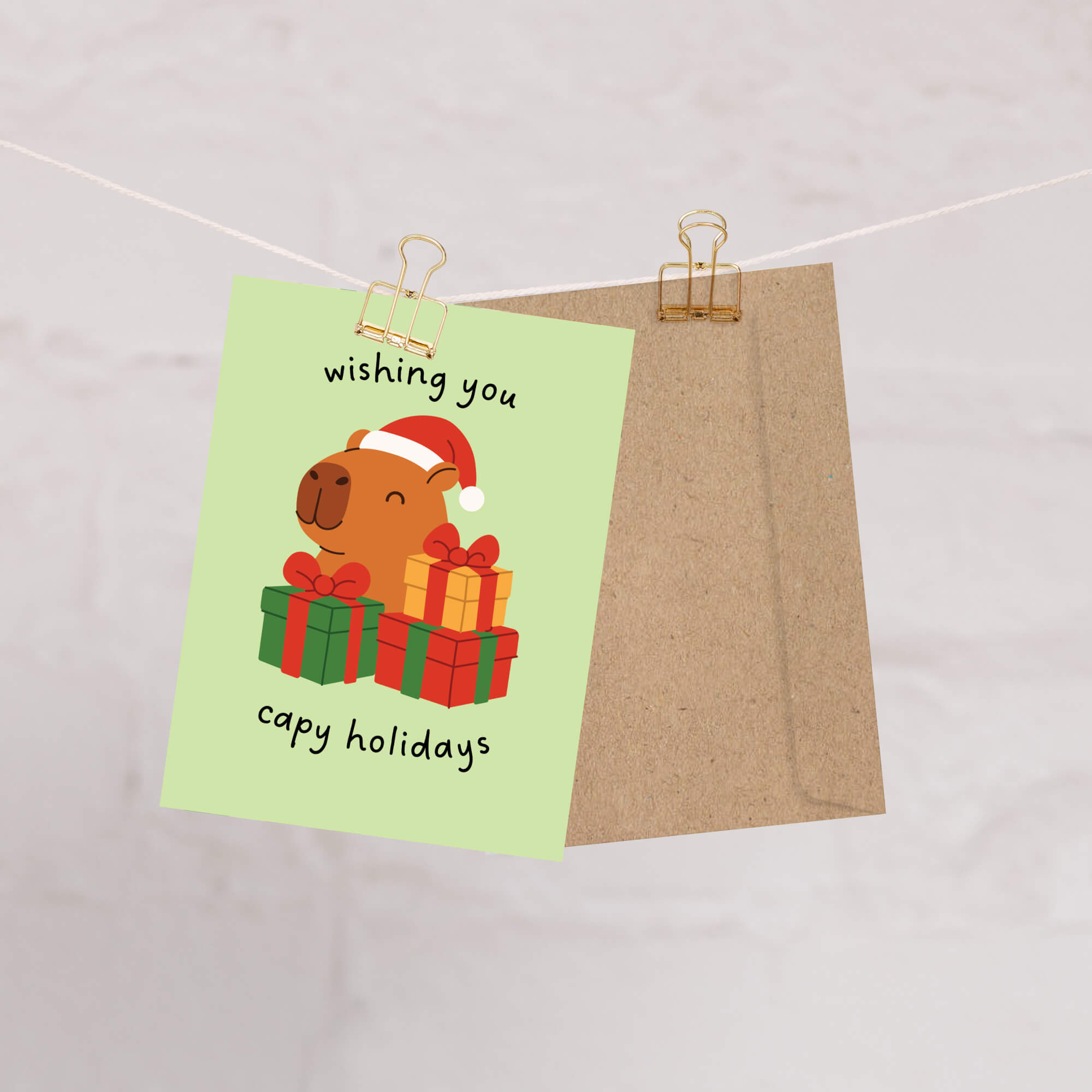 cute unique Christmas card with a capybara wearing a Santa hat and surrounded by presents on a light green background with text pun: wishing you capy holidays