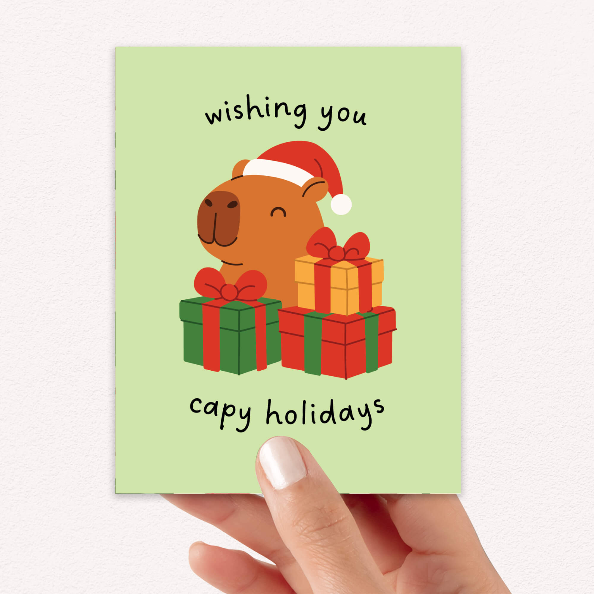 cute unique Christmas card with a capybara wearing a Santa hat and surrounded by presents on a light green background with text pun: wishing you capy holidays