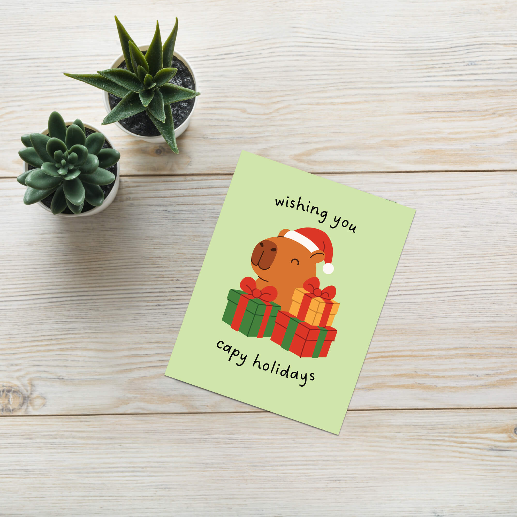 cute unique Christmas card with a capybara wearing a Santa hat and surrounded by presents on a light green background with text pun: wishing you capy holidays