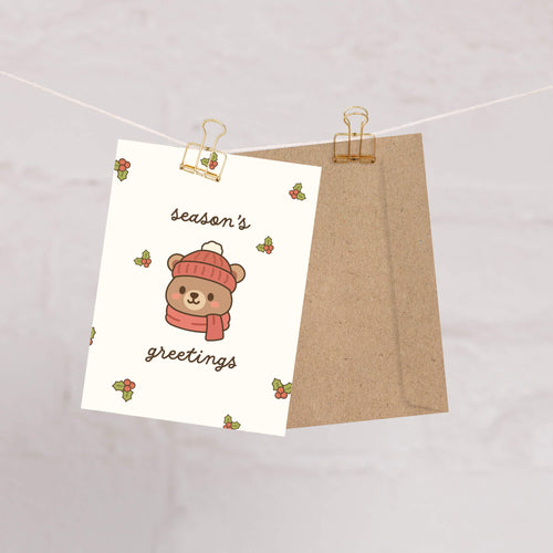 cute Christmas card with a bear wearing a pink hat and scarf, surrounded by holly leaves and berries with text: season's greetings